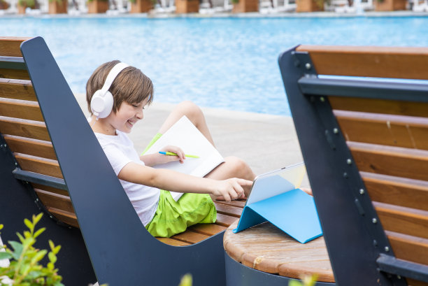 一个小男孩坐在泳池边的躺椅上看在线课程。孩子在学校和世界上任何地方远程学习课程。图片下载