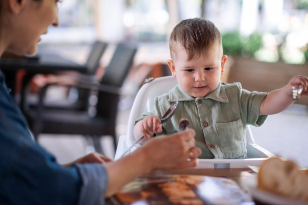 妈妈在自助餐厅给坐在舒适的高脚椅上的小男孩喂健康的食物图片下载
