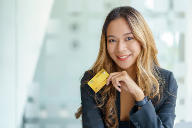 亚洲女商人微笑着看着相机，手里拿着信用卡，在手机或笔记本电脑上进行网上购物。图片下载