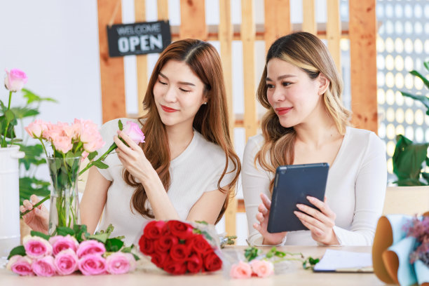 亚洲专业成功的女花商设计师花店老板企业家装饰粉红色的玫瑰花束在玻璃花瓶当同事通过平板电脑在花店工作室在线查看库存图片下载