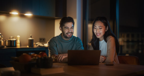 年轻夫妇坐在家里的厨房里微笑着使用笔记本电脑的肖像。订婚夫妇在网上为他们的婚礼准备购物，讨论不同的选择。图片下载