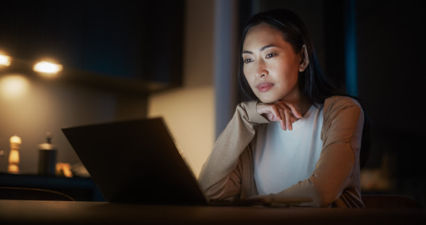 美丽的正宗亚洲女人坐在舒适的厨房里的桌子上，晚上在家使用笔记本电脑。女性在社交媒体上微笑聊天、网购、看新闻图片下载