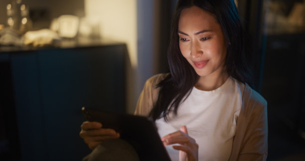 美丽的正宗亚洲女人坐在舒适厨房的桌子上，晚上在家使用数码平板电脑。女性微笑着查看社交媒体、网上购物、观看网络视频。图片下载