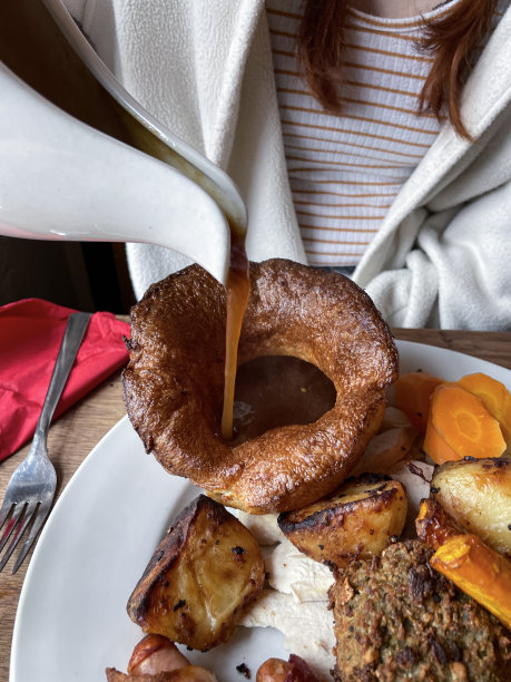特写照片中，一个无法辨认的女人从罐子里倒出肉汁，倒在白色盘子里，盘子里装着周日的烤晚餐、约克郡布丁、胡萝卜、烤土豆、西兰花、裹着毯子的猪、鸡胸肉、鼠尾草和洋葱面包馅图片下载