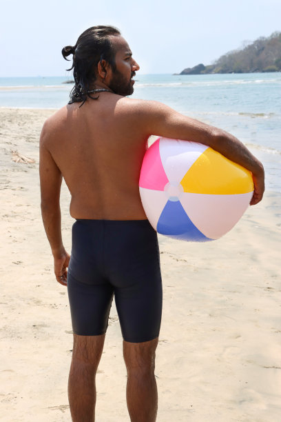 特写照片:赤裸上身的印度男子穿着冲浪短裤站在环礁湖水边的沙滩上，腋下夹着沙滩球和色彩鲜艳的沙滩玩具，看到有棕榈树的林地，侧影图片下载