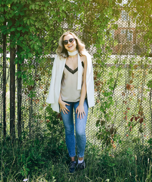 时髦的时髦女孩站在户外常春藤叶墙附近。时尚照片的年轻女性在白色夹克与腰带。金色短发，戴着酷酷的眼镜。夏日生活写真。图片下载