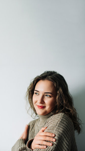 浪漫的妻子热切地期待着丈夫的归来，站在窗前，抱着自己，带着温柔的微笑，深情地凝视着窗外，阳光透过窗户照亮了她的容貌图片下载