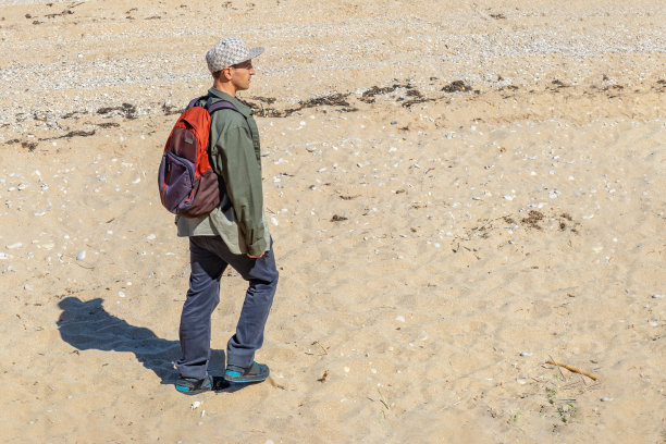 一个穿着牛仔裤、衬衫、棒球帽、背着背包的男人沿着沙滩散步。由于炎热和口渴，人类眯起了眼睛图片下载