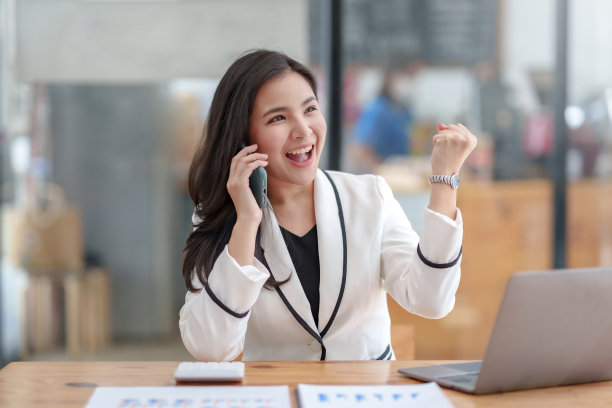 亚洲女商人愉快地坐在笔记本电脑上工作，通过电话联系客户，解释工作，并愉快地微笑。图片下载