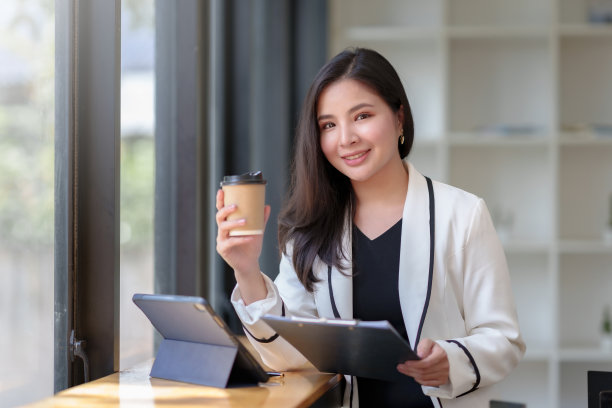 美丽的亚洲女商人坐在她的笔记本电脑上愉快地工作，微笑着喝着咖啡，看着镜头，在办公室里看起来很明亮。图片下载