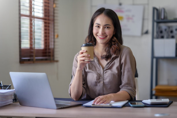 美丽的亚洲女商人坐在她的笔记本电脑上愉快地工作，微笑着喝着咖啡，看着镜头，在办公室里看起来很明亮。图片下载