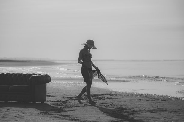 一个模特走在空旷的沙滩上，夕阳下戴着帽子，单色照片。图片下载
