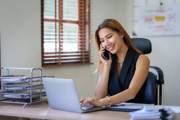 快乐的亚洲女商人微笑着在办公室里和客户讲电话，桌上放着一堆文件。图片下载