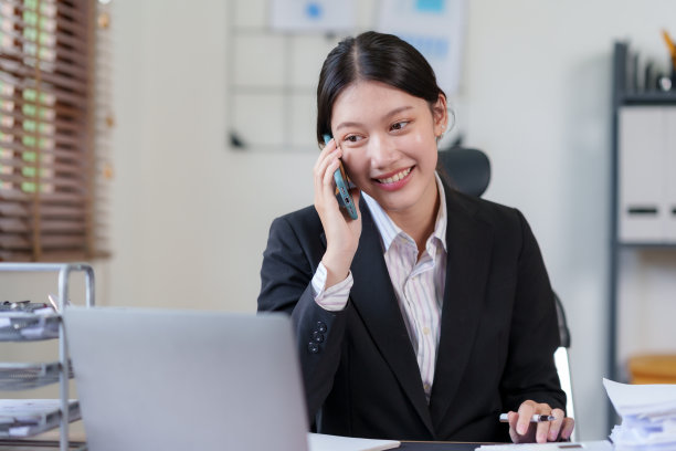 快乐的亚洲女商人微笑着在办公室里和客户讲电话，桌上放着一堆文件。图片下载