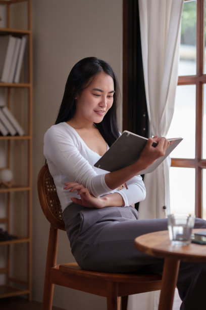 快乐的亚洲女大学生在假期里在客厅里看书和用笔记本电脑工作。亚洲妇女在家看书。图片下载