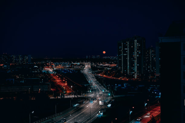 一个美丽的夜晚的城市从一个高大的建筑与天空中的一轮红月亮。明亮的夜晚城市，道路和灯笼，月亮在黑色的天空升起。城市夜生活的概念。图片下载