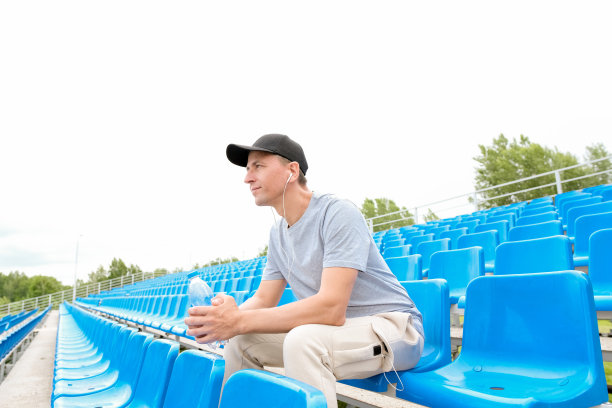 男运动员戴着耳机在体育场外，坐在讲台上听音乐，拿着瓶子喝水图片下载
