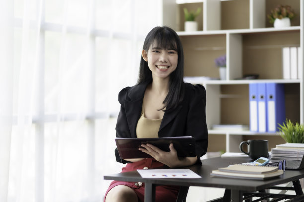 美丽的亚洲女商人在她的笔记本电脑上工作，享受工作，记笔记，回顾工作，开心地微笑着看着镜头。图片下载