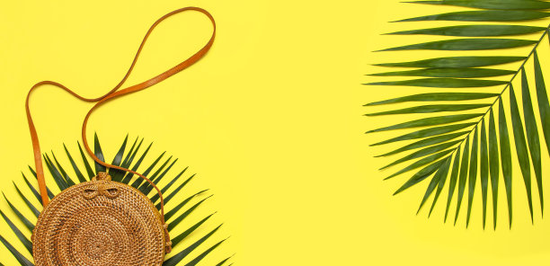 夏季时装平铺。时尚手工自然圆藤袋和热带棕榈叶在黄色的背景。俯视图拷贝空间。来自巴厘岛的时尚竹袋Ecobags。热带的背景图片下载