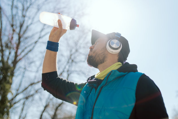 一个戴着帽子戴着耳机的男人，一个运动员，一个穿着无袖夹克的跑步者，他拿着一个水瓶，被太阳晒得很热，春天的天气，一个在公园里跑完马拉松后大汗淋漓的人图片下载