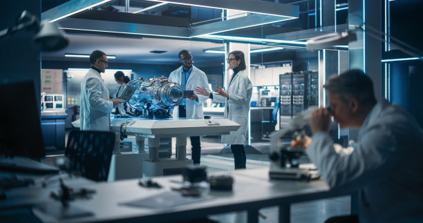 年轻的多元文化工程师团队使用蓝图，平板电脑，笔记本电脑和显微镜来分析和讨论电动涡轮马达的工作原理。高科技研究实验室及设备图片下载