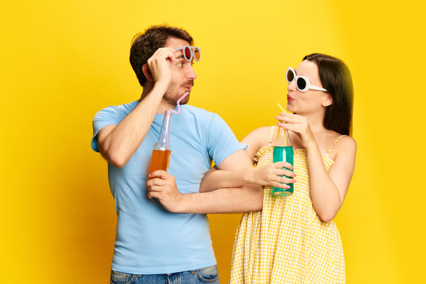 暑假，一对年轻的夫妇，一男一女戴着墨镜，在黄色的摄影棚背景下喝着柠檬水图片下载