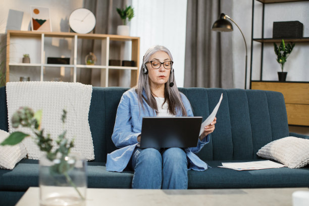 美丽的高级妇女使用耳机和笔记本电脑在家里的客厅视频对话。图片下载