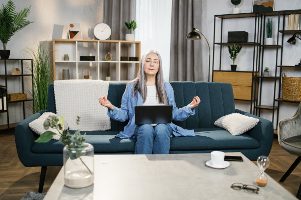 轻松漂亮的花白头发的女士坐在沙发上，膝上放着笔记本电脑，闭着眼睛冥想。图片下载