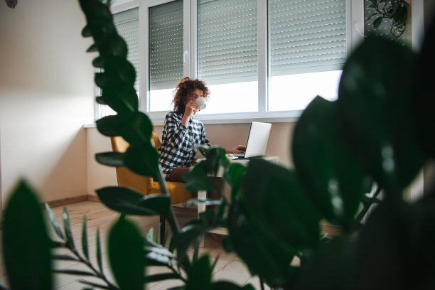 从绿色植物旁看一位卷发女人站在笔记本电脑前喝咖啡，等待视频会议开始。居家办公。新冠疫情。图片下载