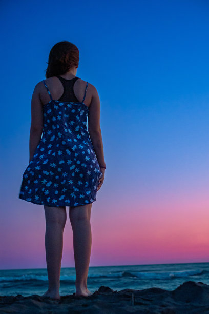 女孩站在靠近水边的沙滩上，看着日落后平静的海浪图片下载