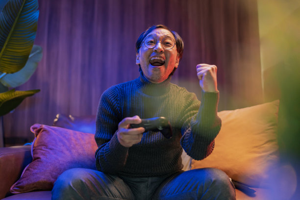 退出愉快的亚洲日本中年成年人晚上独自在家玩电子游戏，在黑暗中坐在沙发上的游戏控制器，亚洲男性反应享受自己的胜利，在晚上的时候举起游戏手，在天空中喊着胜利者的问候图片下载