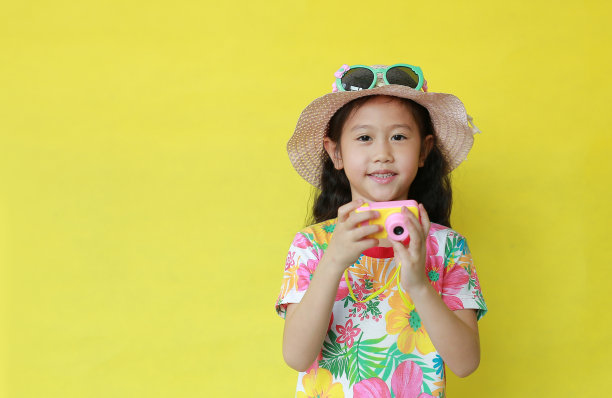 小女孩手里拿着玩具相机。亚洲孩子的肖像在花图案夏季连衣裙和帽子与太阳镜孤立的黄色与复制空间。时尚的假日和夏季的概念图片下载