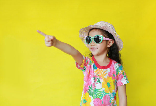 快乐的亚洲小女孩穿着花图案的夏季连衣裙和帽子，太阳镜指向黄色背景的复制空间。假日和夏季时尚概念。图片下载