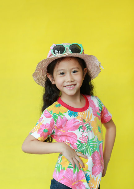 可爱的亚洲小女孩微笑着穿着花图案的夏装，戴着帽子和太阳镜，在黄色的背景下摆姿势。假日和夏季时尚概念图片下载