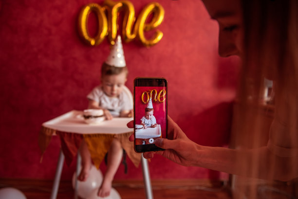 母亲用手机给儿子拍照。一岁大的男孩戴着派对帽，拿着生日蛋糕图片下载