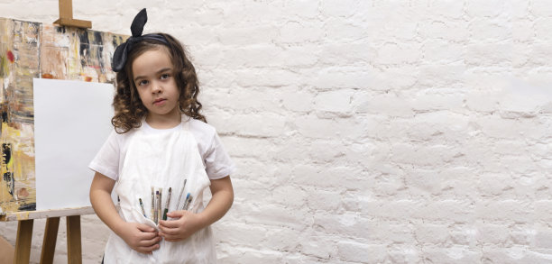 才华横溢的小艺术家与白色围裙和画笔附近的画架。艺术学校工作坊。副本的空间。图片下载