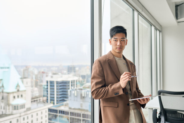 年轻的亚洲商人拿着平板电脑看着窗户思考。图片下载