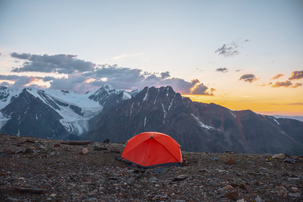 风景优美的高山景观，帐篷位于非常高的海拔，在橙色的黎明天空中可以看到大山。鲜艳的橙色帐篷，在多云的天空下，在日落的颜色下，可以看到令人敬畏的高山。图片下载