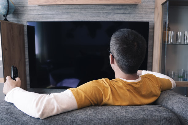 身穿黄色t恤的男性观众对着黑色的电视屏幕。年轻英俊的男人在家里的沙发上看电视图片下载