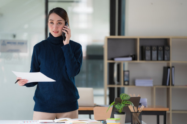 一名亚洲女商人在办公室工作时手持文件报告并使用手机的形象。图片下载
