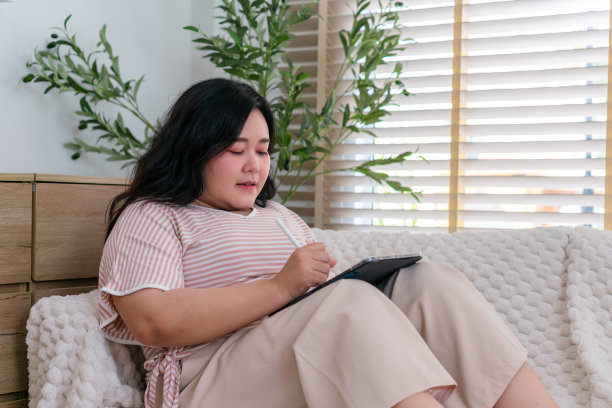 身着便服、头戴红色耳机的年轻女子在客厅的沙发上半坐半跳，心情愉快地使用着平板电脑。图片下载