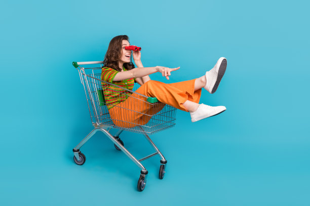 完全尺寸的照片欣喜若狂可爱的女人坐在购物车里看在双筒望远镜指导空空间孤立的蓝色背景图片下载