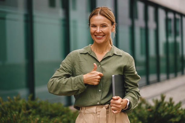 拿着笔记本电脑的女商人站在现代办公楼的背景上，竖起大拇指图片下载