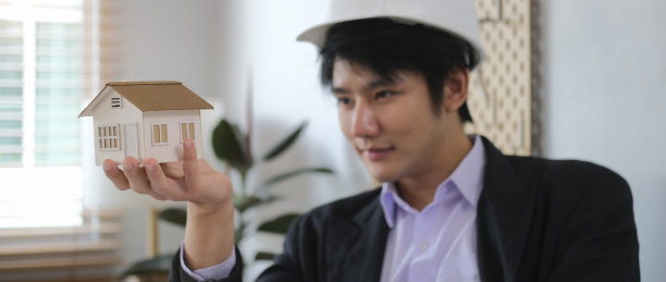 英俊的男建筑师穿着正式的西装，戴着安全帽，拿着房子的小模型。图片下载