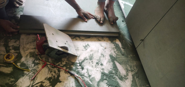 正在进行中的工作——一名木匠在建筑工地用圆形电锯工作， 与绿色的木板一起，凌乱地散落着锯末，电线和机器附近的木板上有一个人的赤脚和手图片下载