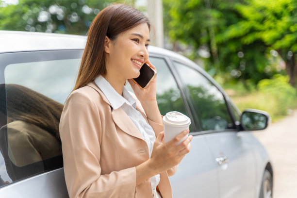 一位年轻的职业女性，面带微笑，手里拿着一杯外带咖啡，靠在一辆汽车上讲着电话图片下载