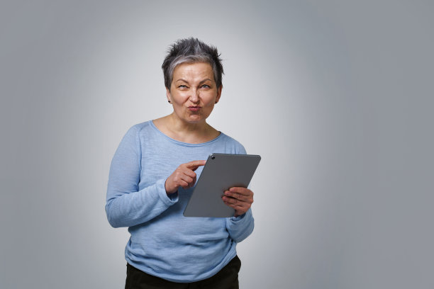 俏皮的灰色头发成熟女性与数字平板电脑工作或检查社交媒体。漂亮的女人在50年代蓝色衬衫孤立在白色背景。老年人与科技图片下载