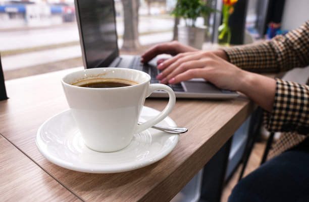 一杯咖啡放在咖啡馆的桌子上，背景是一个在笔记本电脑前工作的女孩的模糊的手。在线工作。特写镜头。有选择性的重点。图片下载