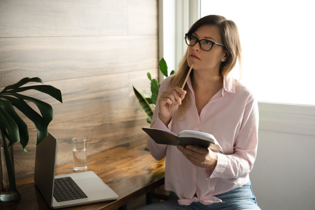 有魅力、有思想的中年妇女，戴着眼镜，留着长长的直发，在家里办公室用笔记本电脑工作，笔记本电脑放在桌子上。远程工作概念。本空间图片下载