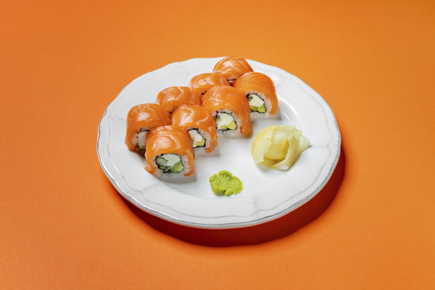 费城卷，鲑鱼，米饭和费城奶酪馅黄瓜。寿司放在一个白色的圆形陶瓷盘子上，盘子上有图案。盘子立在桔黄色的纸底上。图片下载